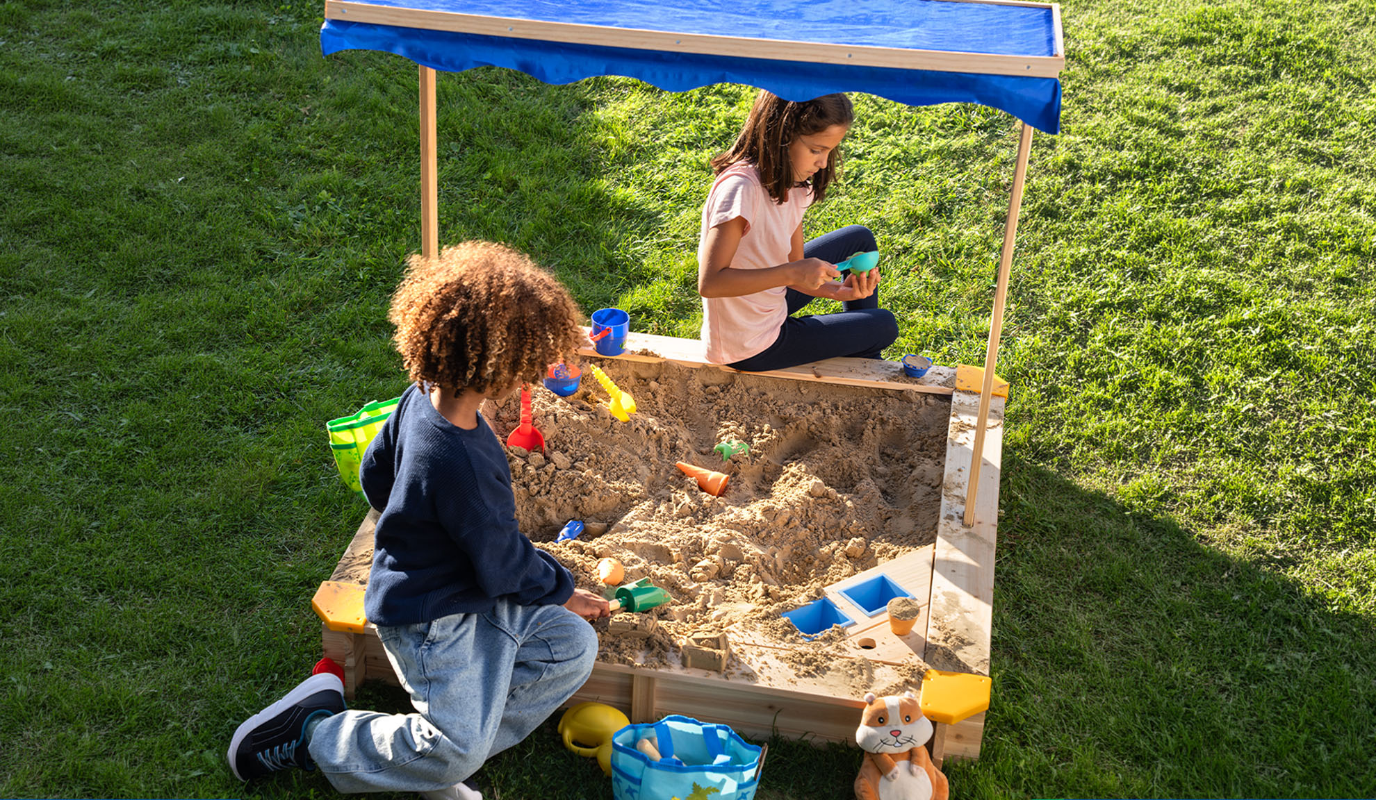 Zwei Kinder spielen in einem Sandkasten mit blauem Dach und Sandspielzeug.