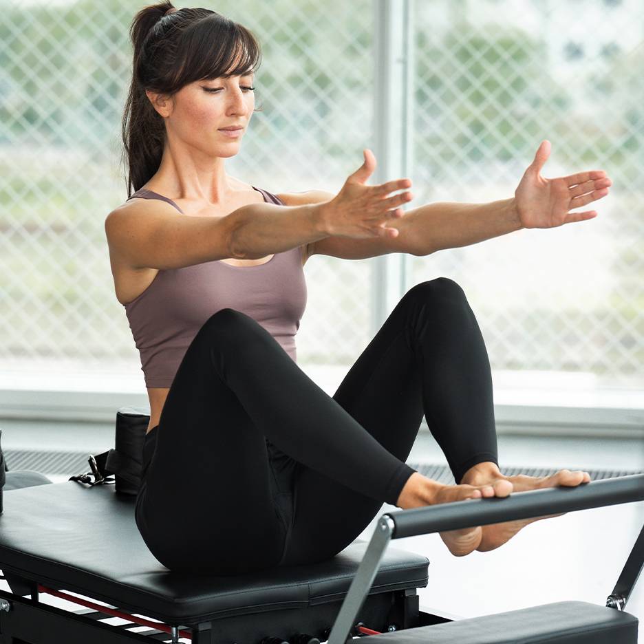 Frau macht Pilates auf einem Reformer, trägt Top und schwarze Leggings.