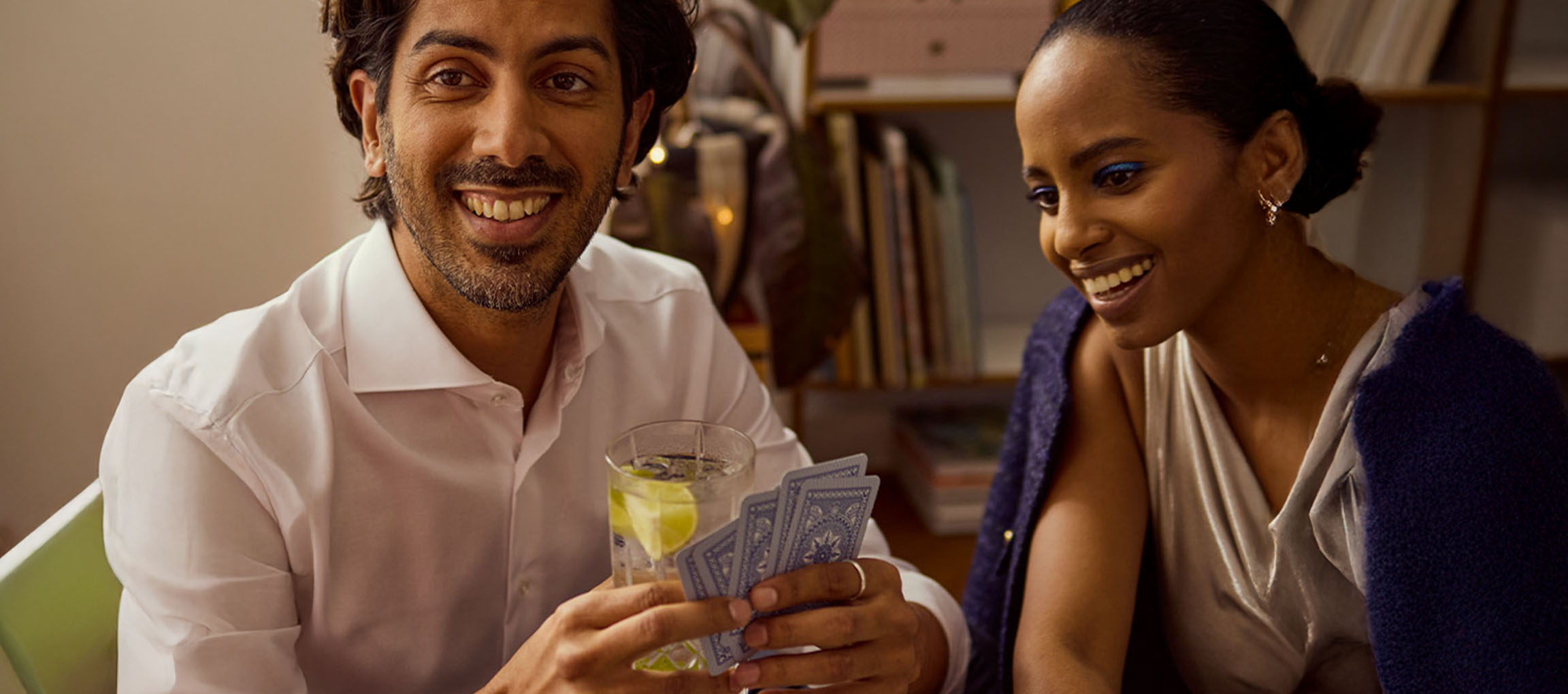 Ein lächelnder Mann und eine Frau spielen Karten, der Mann hält ein Glas.
