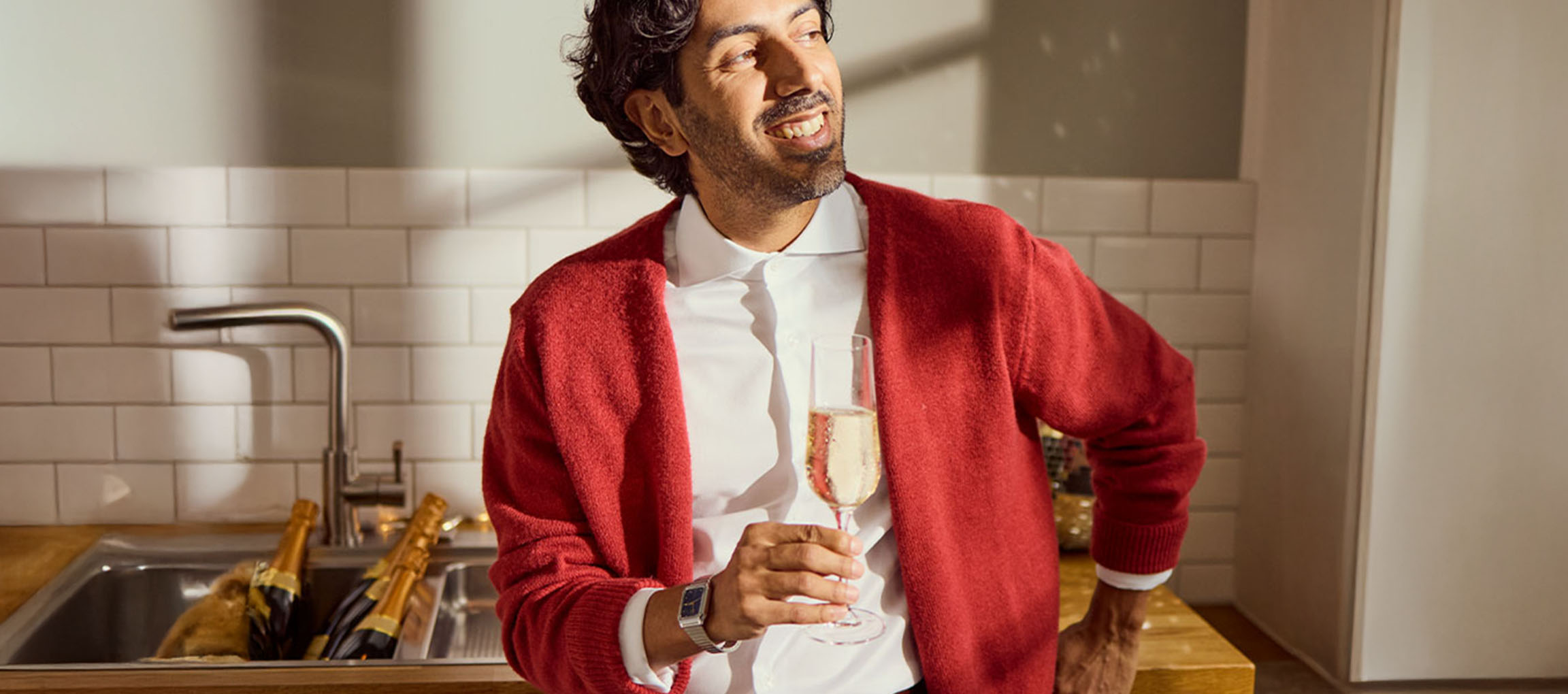 Lächelnder Mann in weißem Hemd und roter Strickjacke, hält ein Glas Champagner.