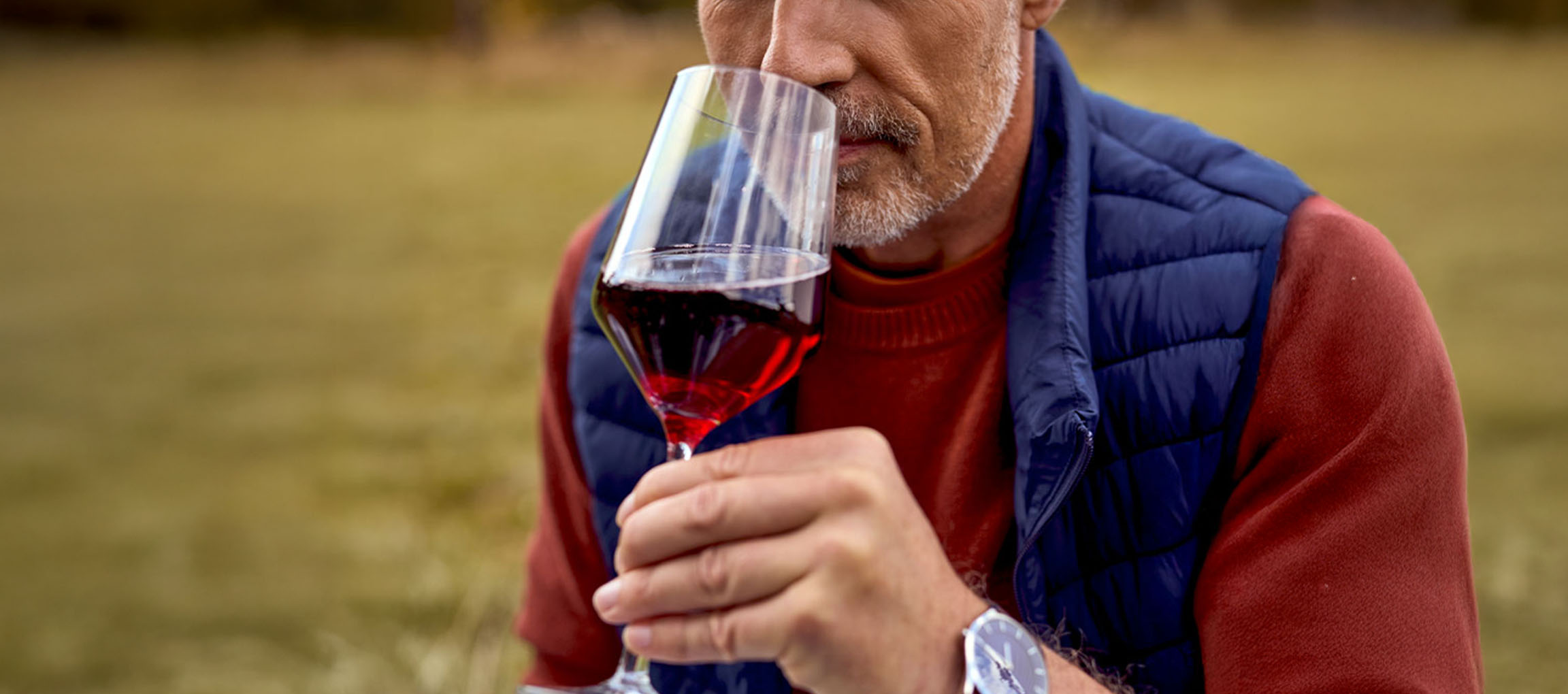 Bärtiger Mann riecht an Rotwein aus einem Glas, trägt Weste und Pullover.