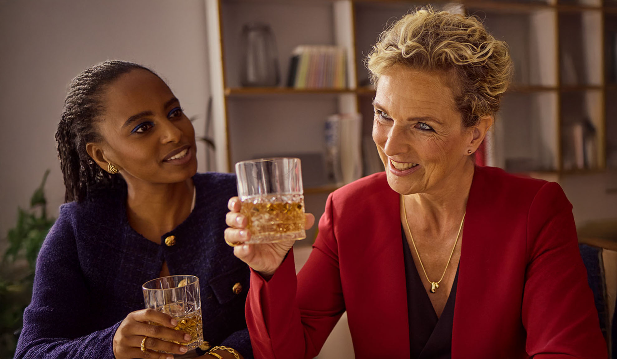 Zwei lächelnde Frauen halten Gläser mit Getränken, eine in roter Jacke, die andere in marineblauer Jacke.