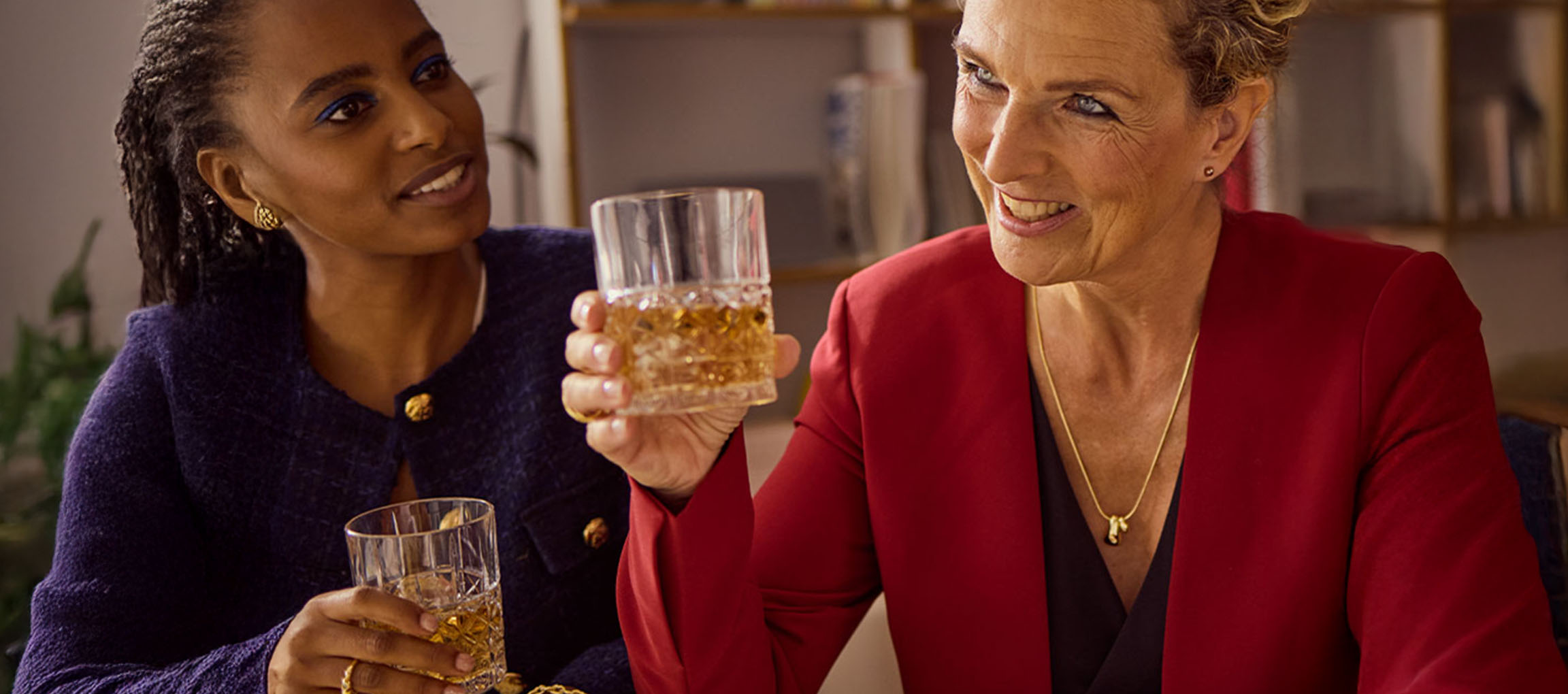 Zwei Frauen in Blazern halten Gläser mit bernsteinfarbener Flüssigkeit, lächeln und schauen sich an.