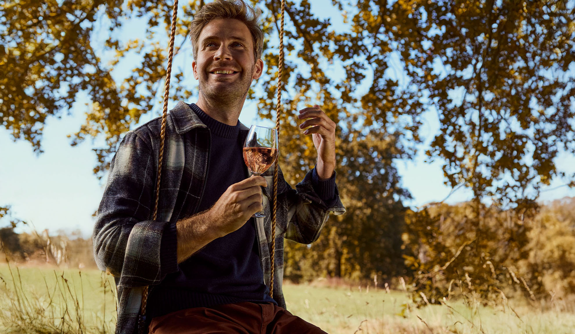 Lächelnder Mann auf einer Schaukel, der ein Glas Roséwein im Freien hält.
