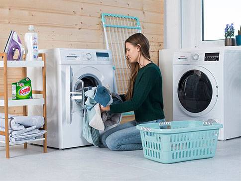Waschmaschine, Trockner und Wäschekorb; Frau legt Wäsche in die Waschmaschine.