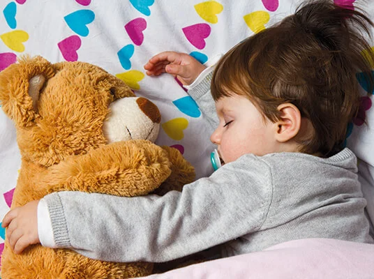 Baby schläft mit Kuscheltier in einem Bett mit Herzmuster.