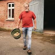 Ein Mann trägt einen Korb mit Kartoffeln vor einem alten Bauernhaus.