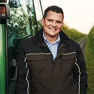 Ein lächelnder Landwirt steht vor einem grünen Traktor.