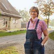 Eine Frau in Jeans-Latzhosen steht lächelnd vor einem Fachwerkhaus.