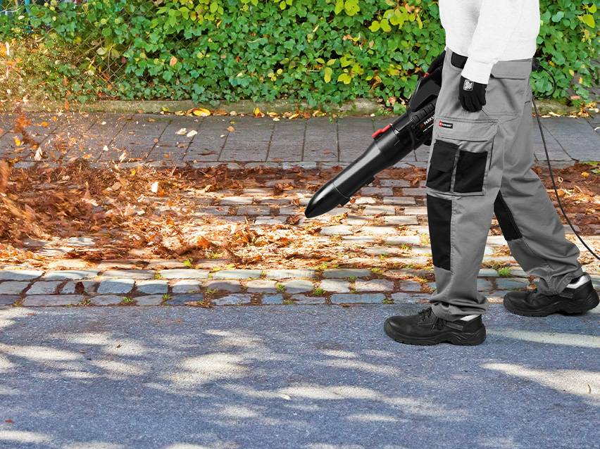 Eine Person bläst mit einem Laubbläser Blätter von einem gepflasterten Weg.
