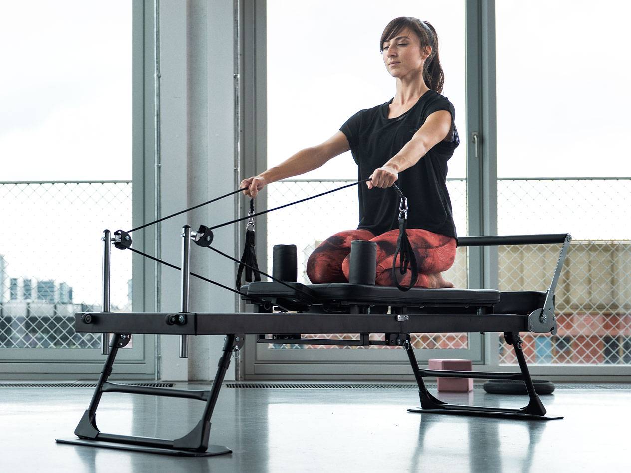 Frau macht Pilates auf einem Reformer vor großen Fenstern.