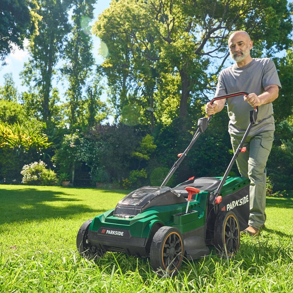 Mann mäht Rasen mit grünem Rasenmäher in sonnigem Garten.