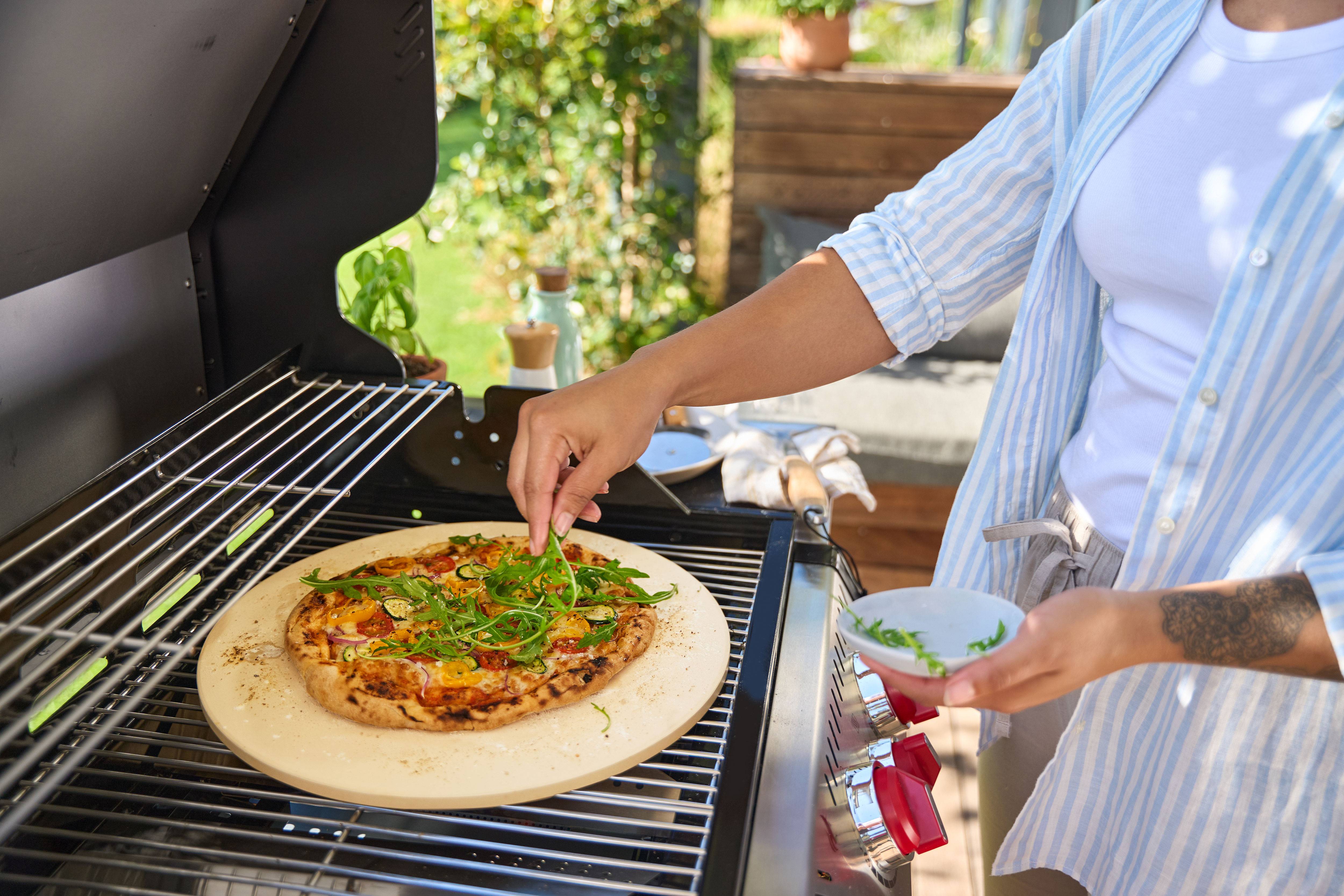 Person belegt Pizza auf Pizzastein im Gasgrill mit Rucola.