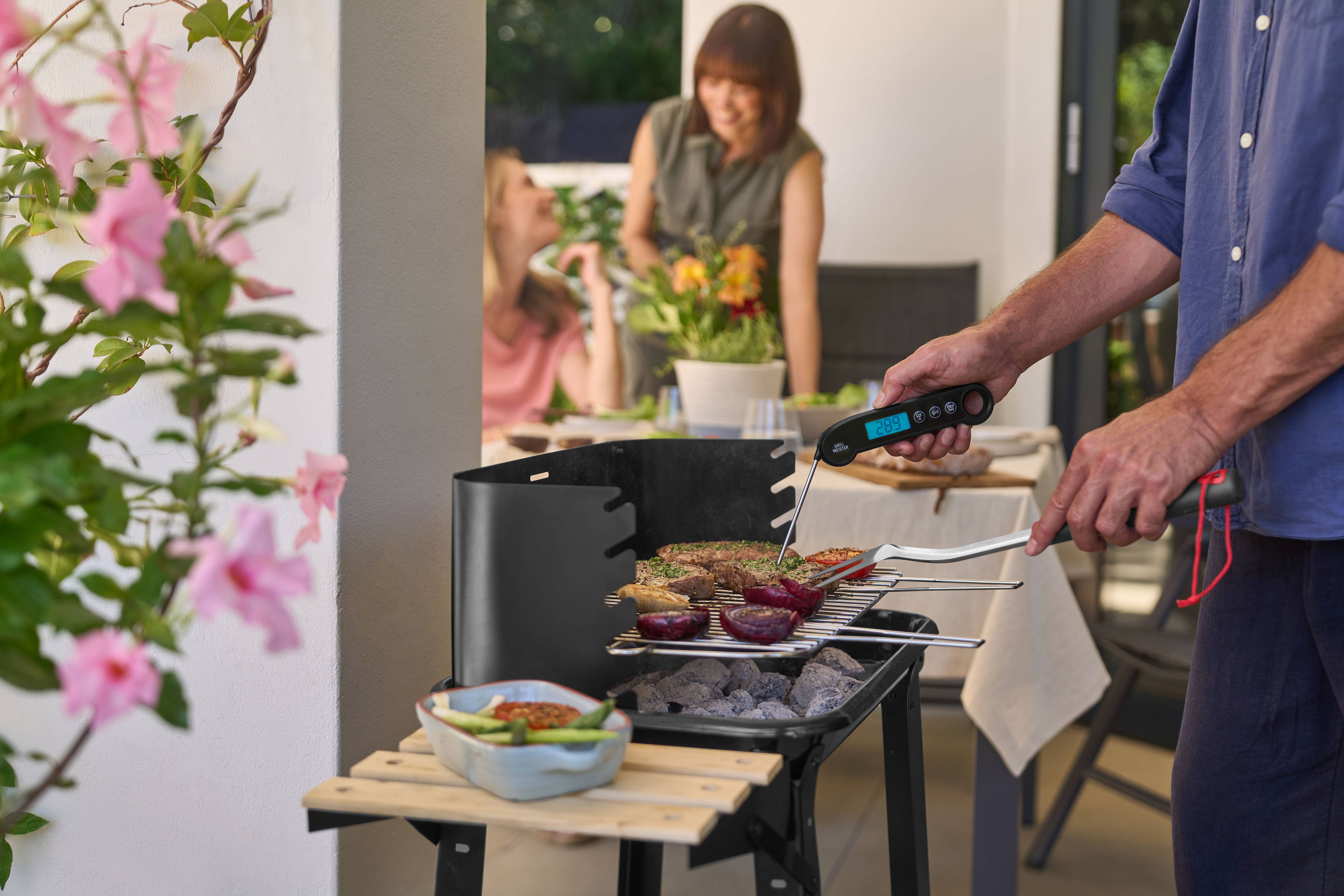 Mann grillt Fleisch und Gemüse mit einem Fleischthermometer, Frauen im Hintergrund.