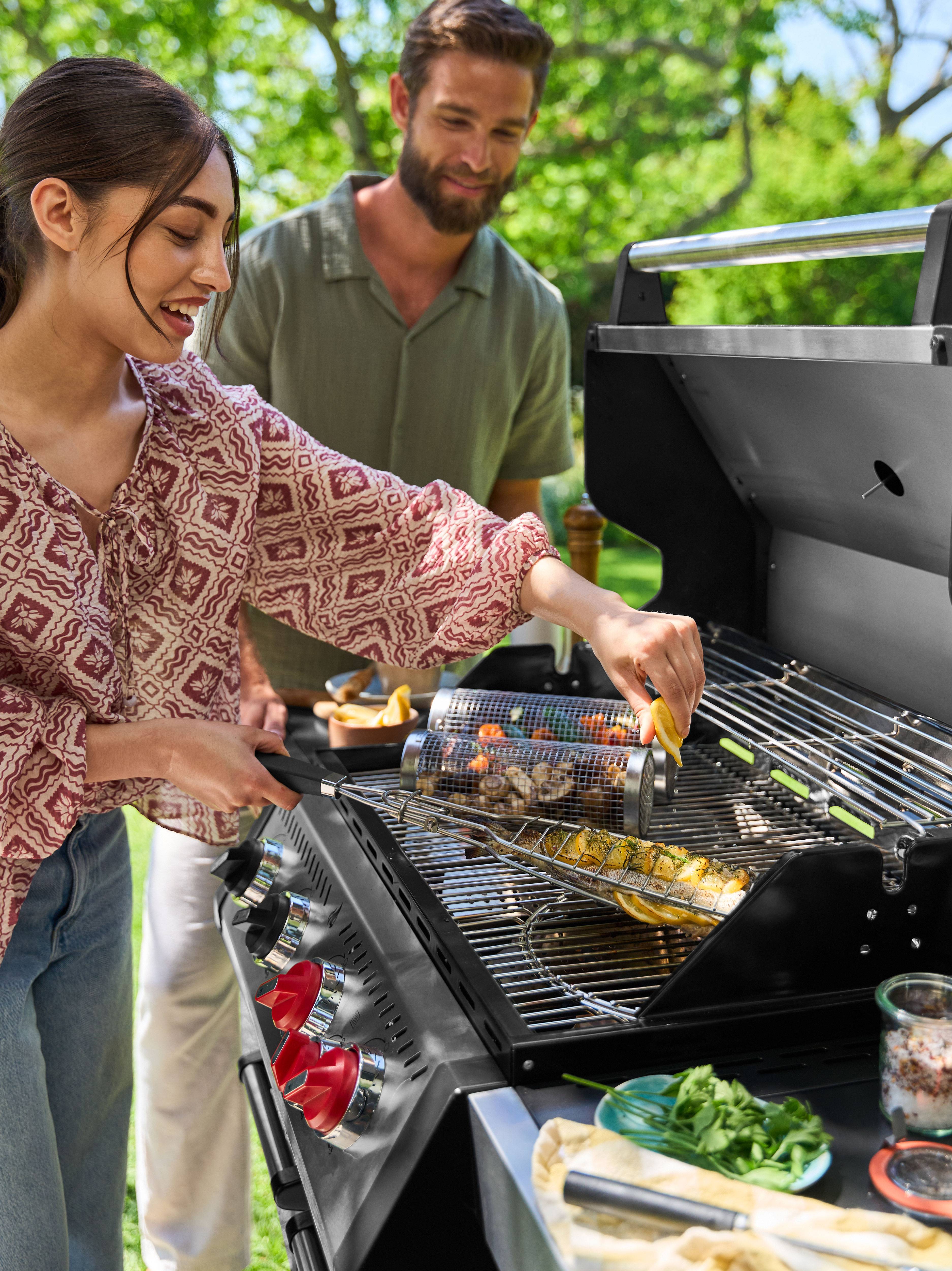 Lächelnde Frau grillt Fisch und Gemüse auf Gasgrill, Mann schaut von hinten zu.