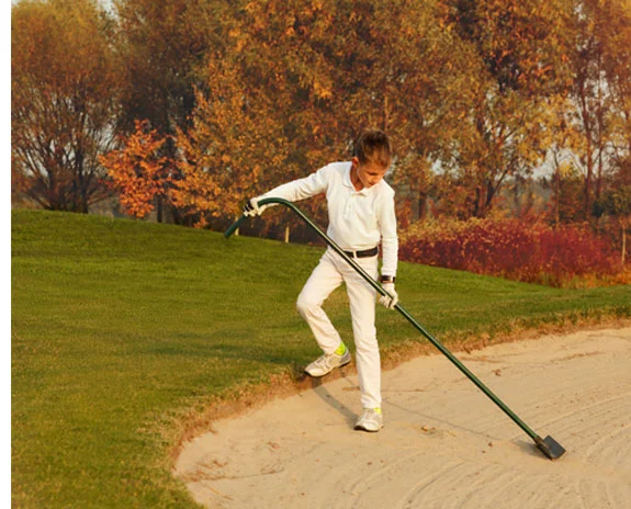 Kind harkt im Herbst auf einem Golfplatz Sand.