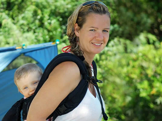 Frau trägt Baby in Tragehilfe in der Natur, neben Zelt.