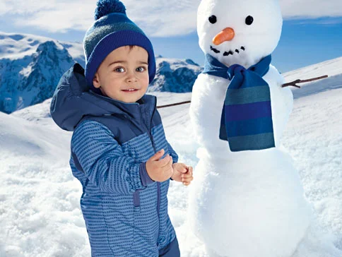 Kleinkind in blauem Schneeoverall und Mütze neben einem Schneemann.
