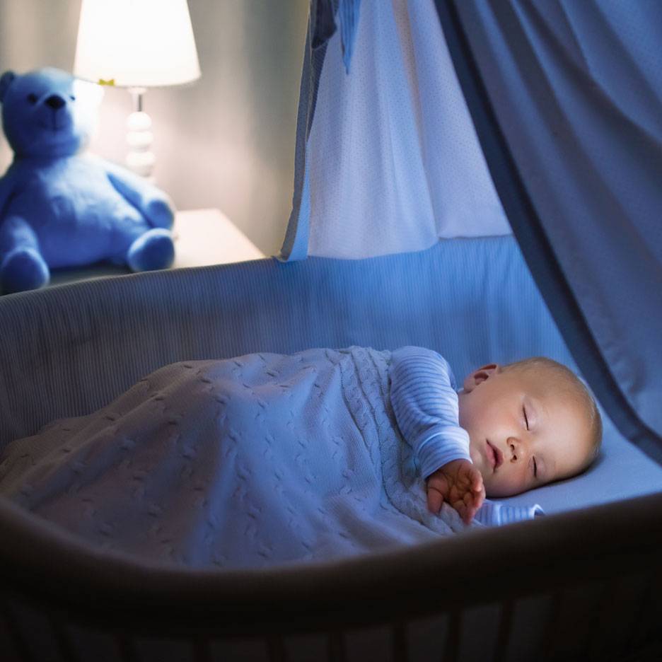 Schlafendes Baby im Kinderbett mit blauem Teddybär und Nachtlicht.