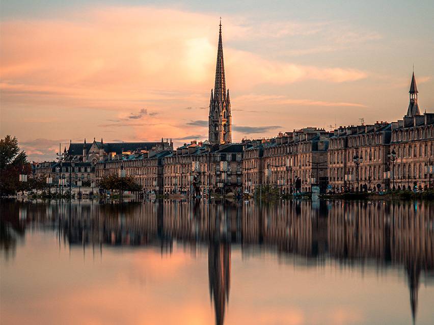 Stadtbild von Bordeaux bei Sonnenuntergang mit einer Kirche und ihrer Spiegelung im Wasser.