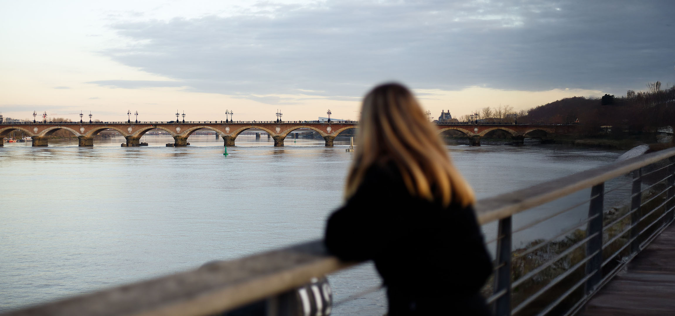 Blick auf eine Bogenbrücke und einen Fluss, mit einer Person mit langen Haaren im Vordergrund.