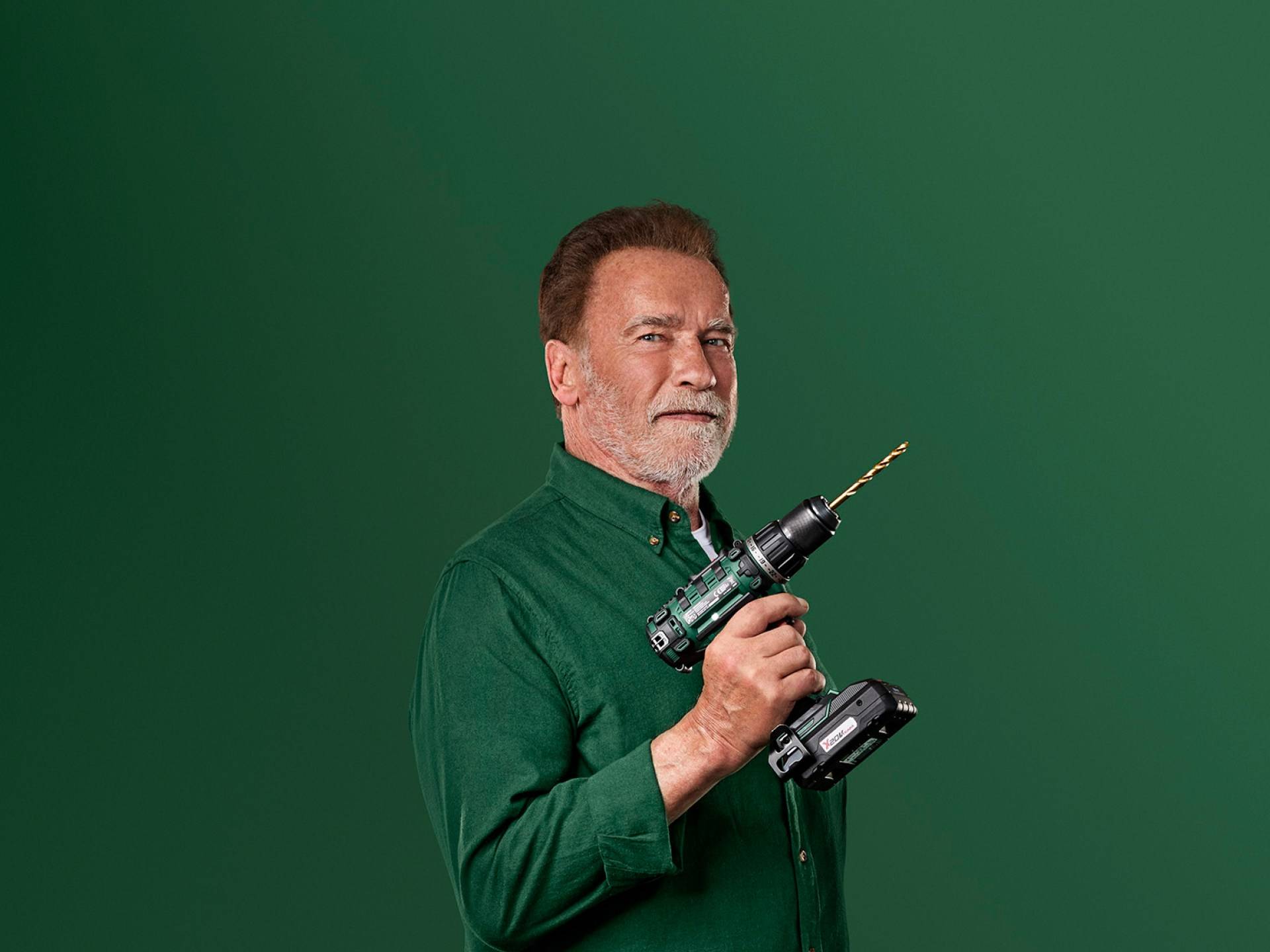 Arnold Schwarzenegger hält einen grünen Akkuschrauber vor grünem Hintergrund.