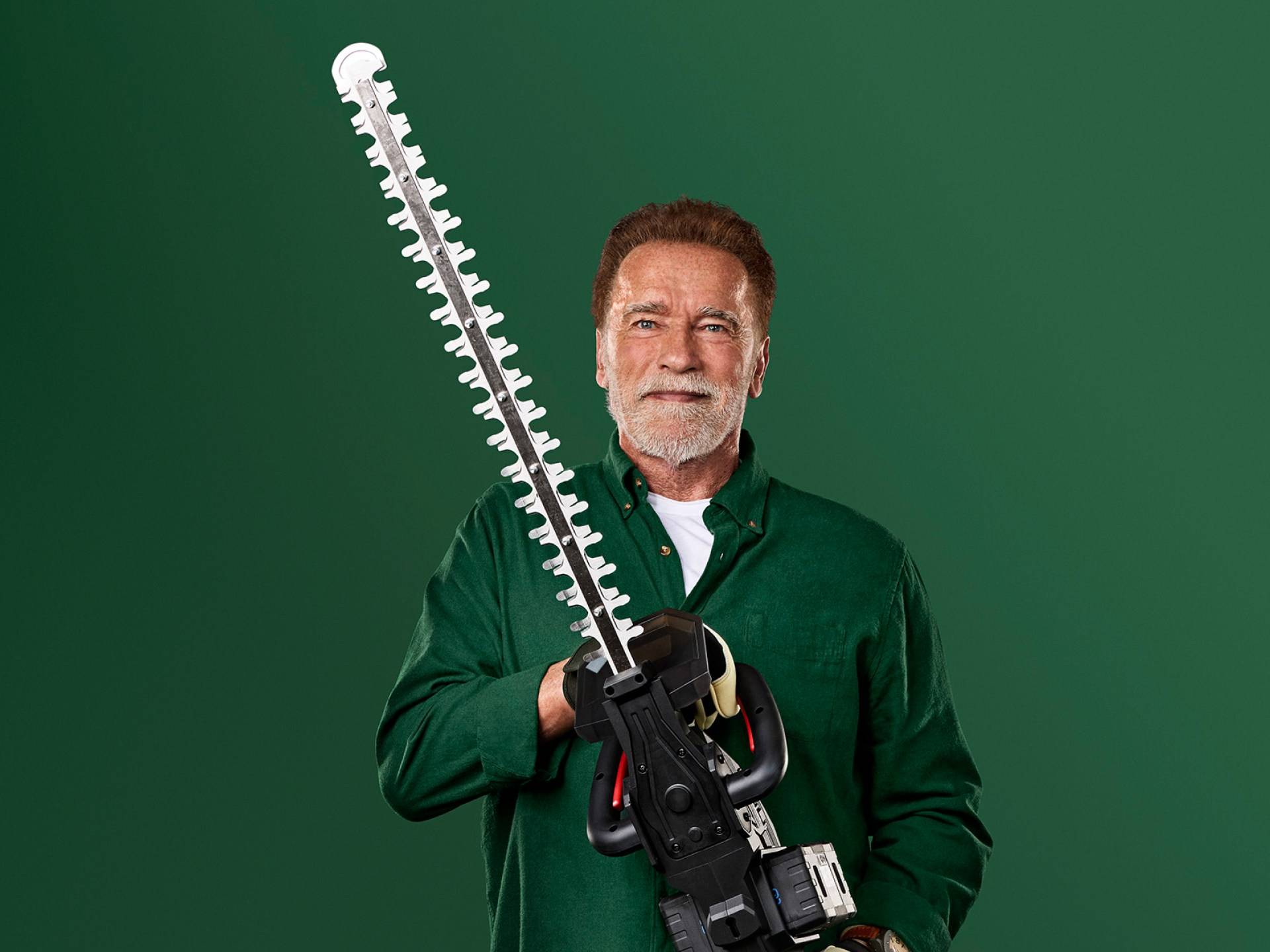 Arnold Schwarzenegger hält eine große Heckenschere vor grünem Hintergrund.