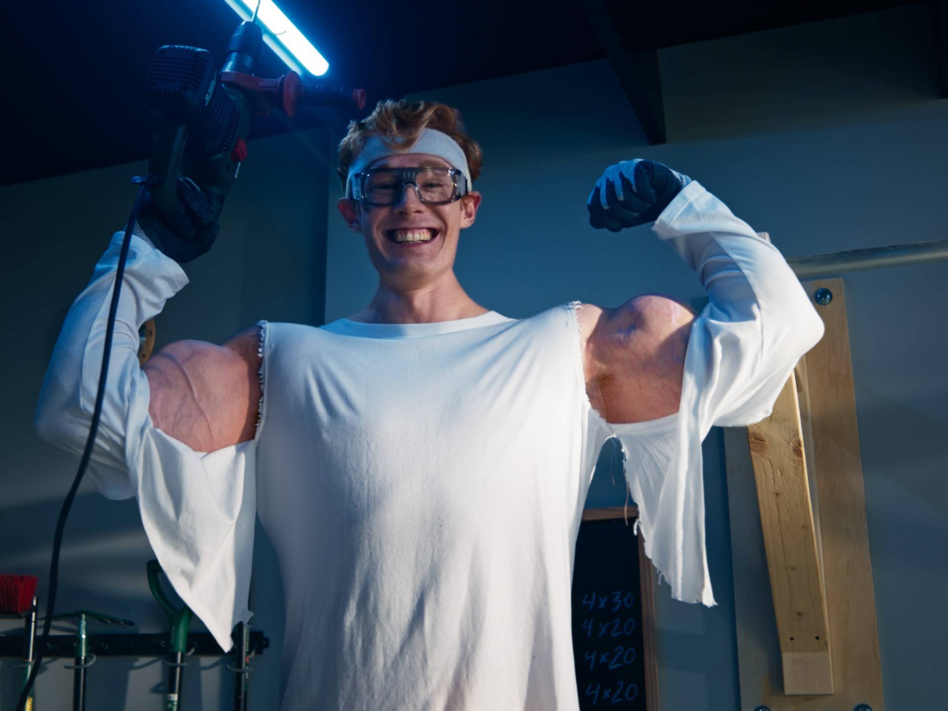 Mann mit Werkzeugen und überdimensionierten Muskeln lächelt in einer Werkstatt.