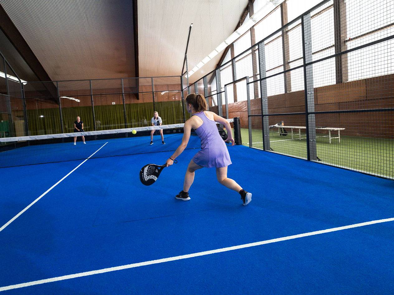 Frau spielt Padel auf einem blauen Platz, mit anderen Spielern im Hintergrund.