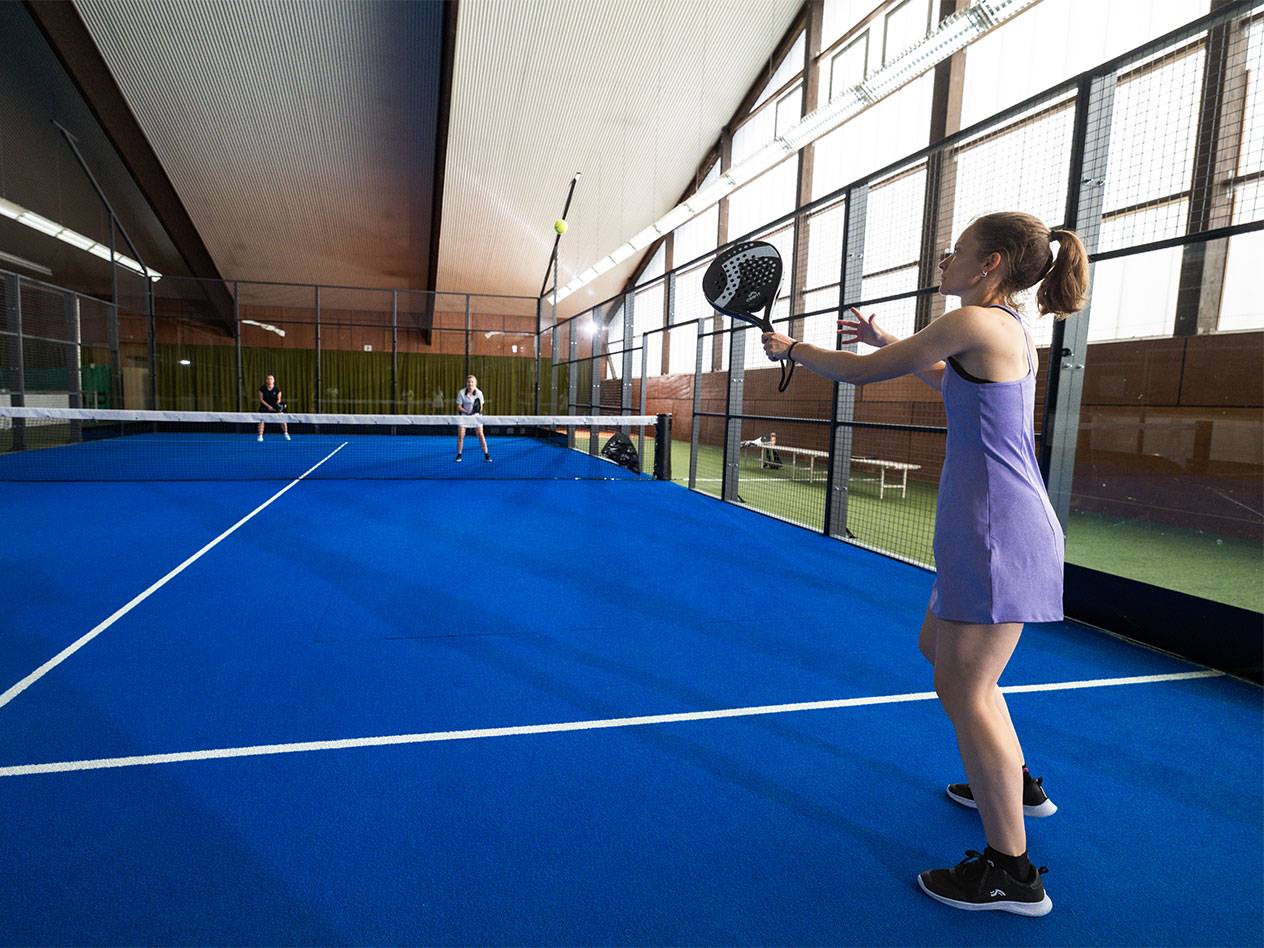 Frau in lila Kleid spielt Padel auf blauem Platz, andere Spieler im Hintergrund.
