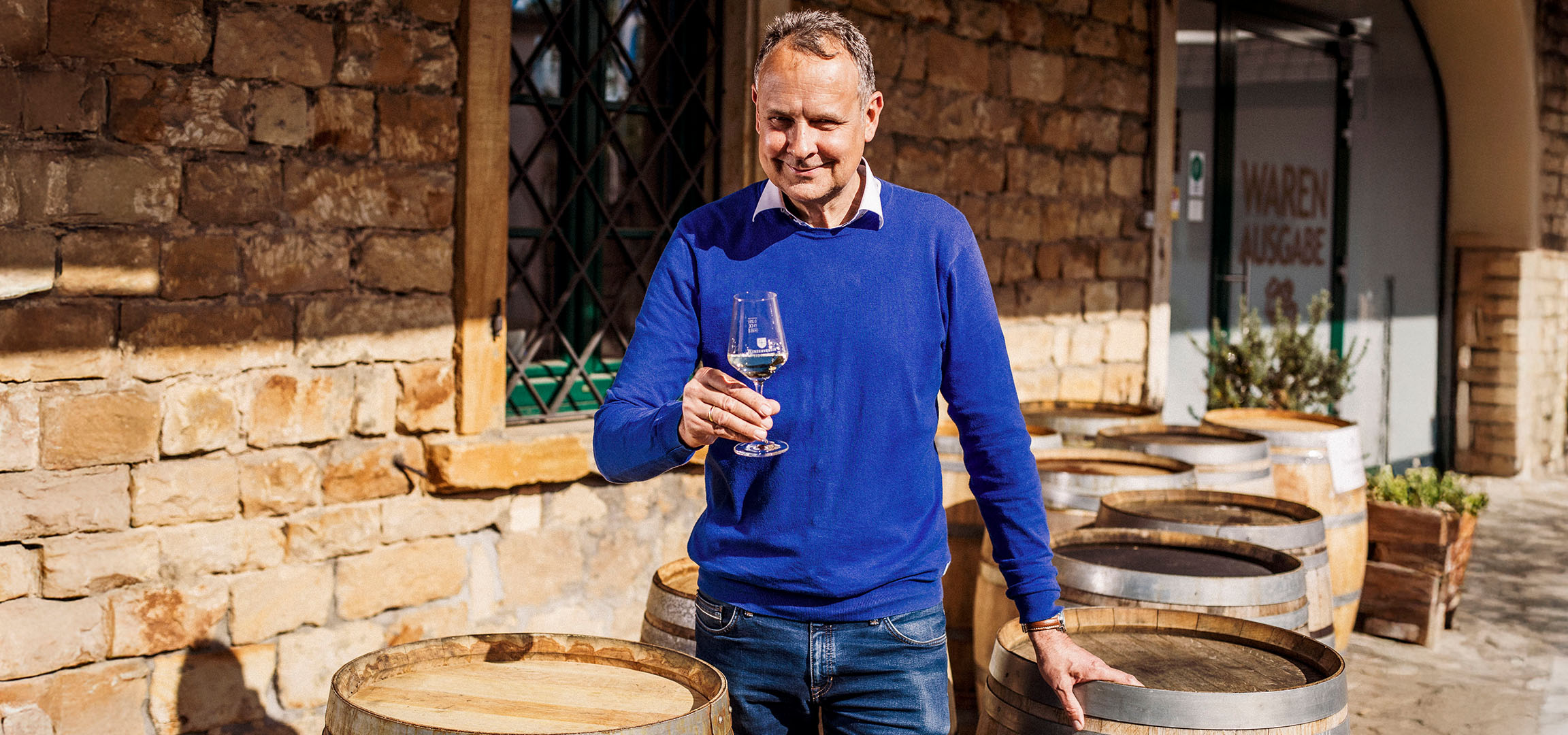 Lächelnder Mann in blauem Pullover hält ein Glas Weißwein vor einer Steinmauer.