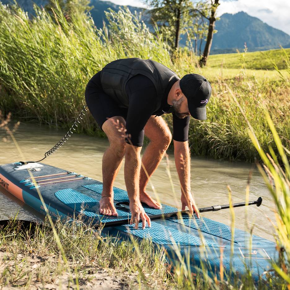 Mann in Neoprenanzug und Schwimmweste bereitet sich auf Stand-Up-Paddling auf einem Fluss vor.