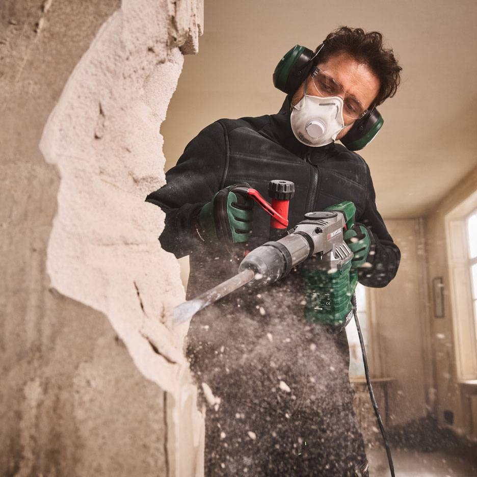 Mann mit Abbruchhammer und Schutzausrüstung reißt eine Wand ab.