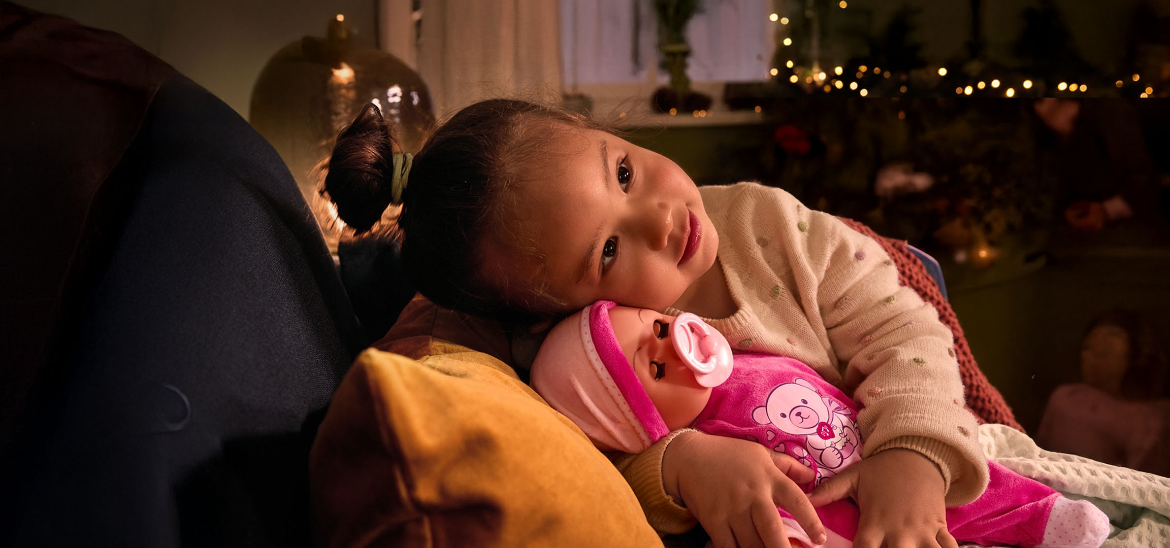 Mädchen mit Puppe und Schnuller, gemütlich auf einem Kissen.