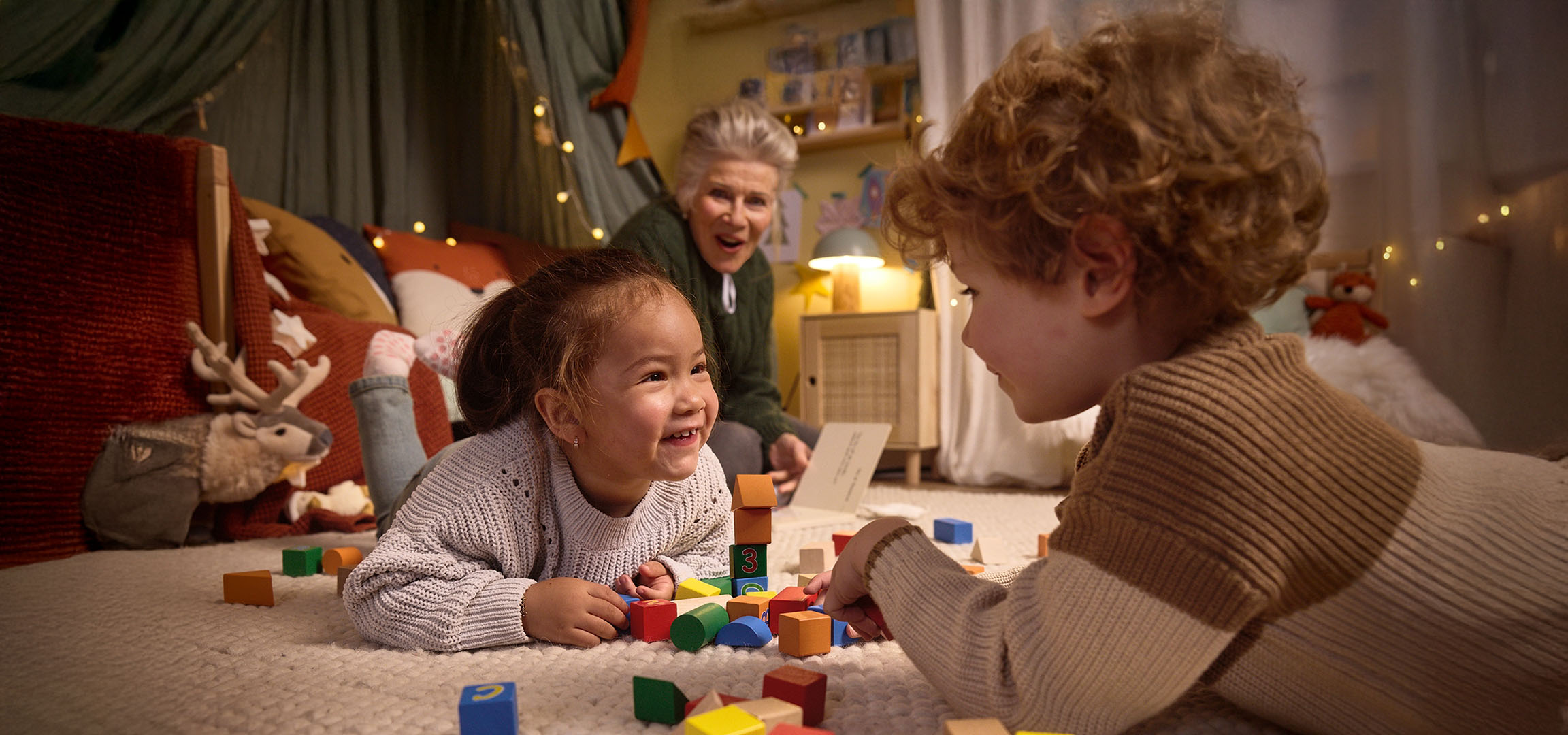Zwei Kinder spielen mit bunten Bauklötzen auf dem Boden, ein Erwachsener schaut zu.