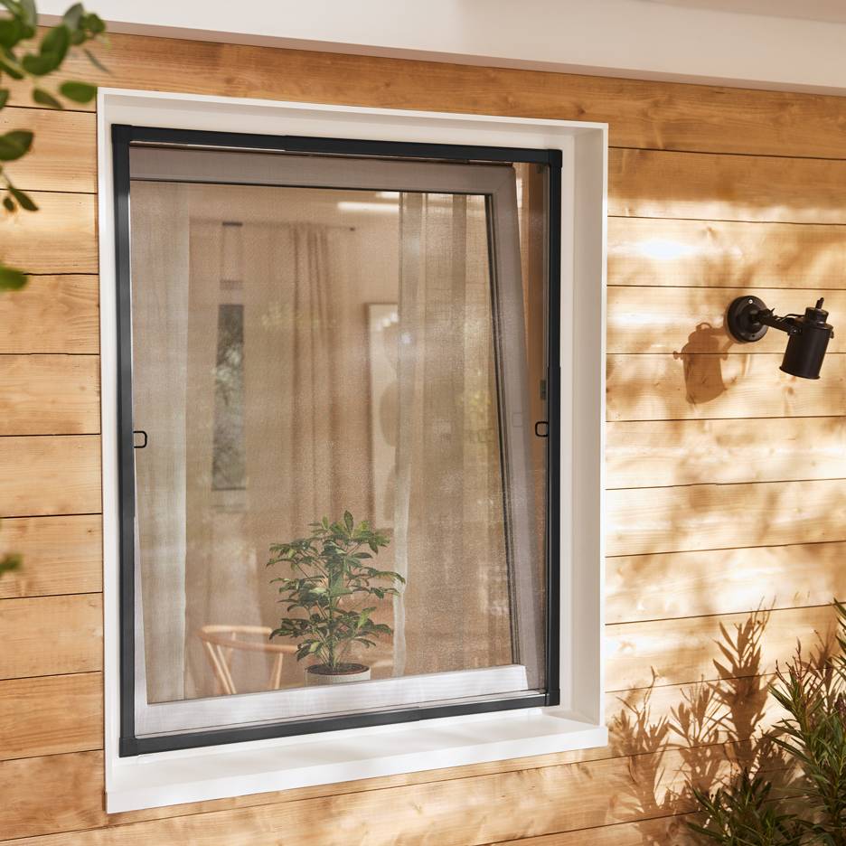 Fenster mit ausziehbarem Insektenschutzgitter an einer Holzfassade, mit einer Pflanze darin.