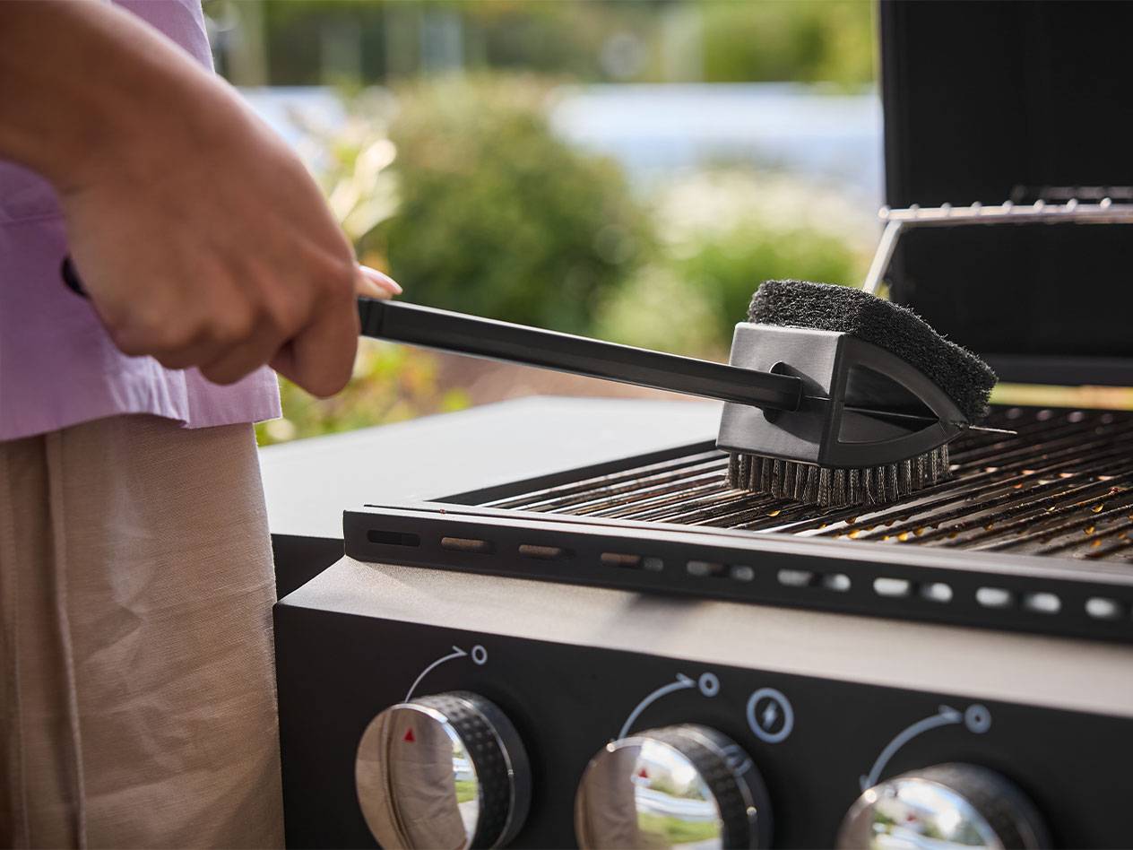 Person reinigt einen Grill mit einer langstieligen Bürste.