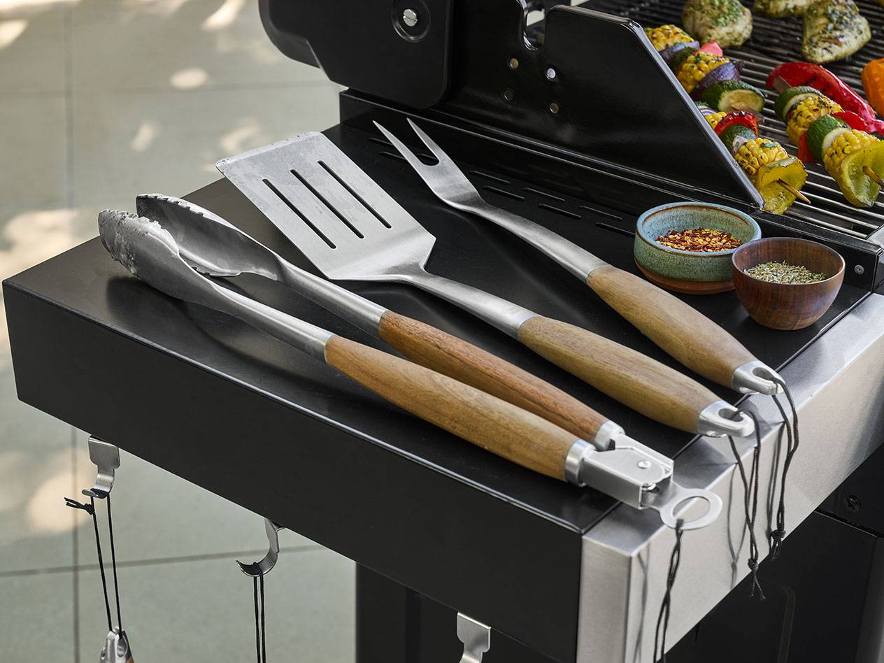 Grillbesteck mit Holzgriffen und Gewürze neben gegrillten Gemüsespießen.