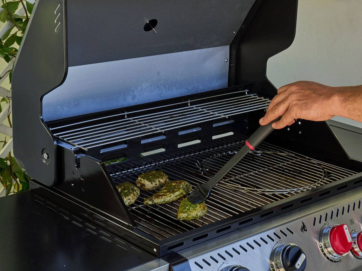 Eine Person bestreicht gewürzte Hähnchenstücke auf einem Gasgrill mit einem Pinsel.