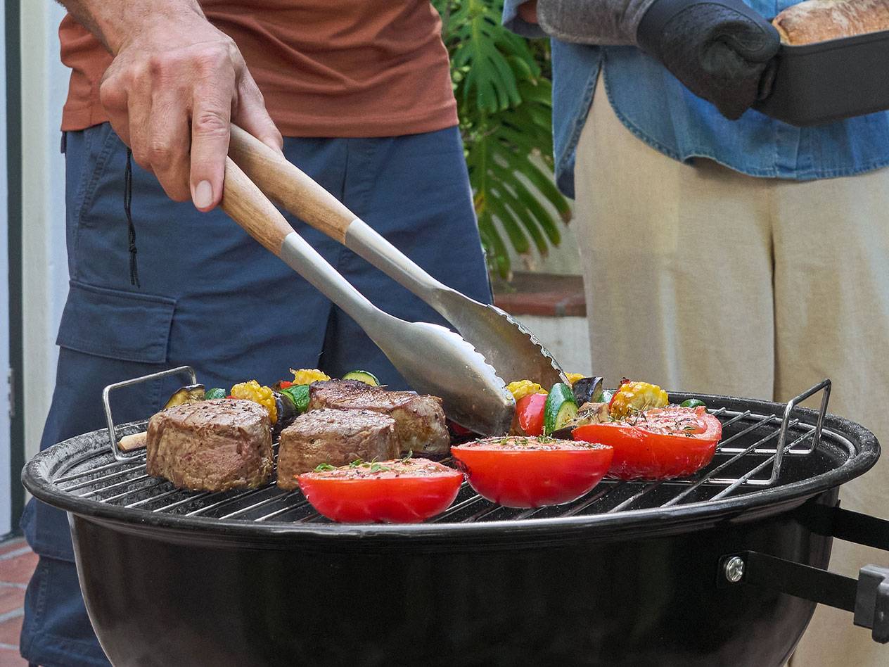 Mann grillt Fleisch, Gemüse und Tomaten, Frau hält Brot.