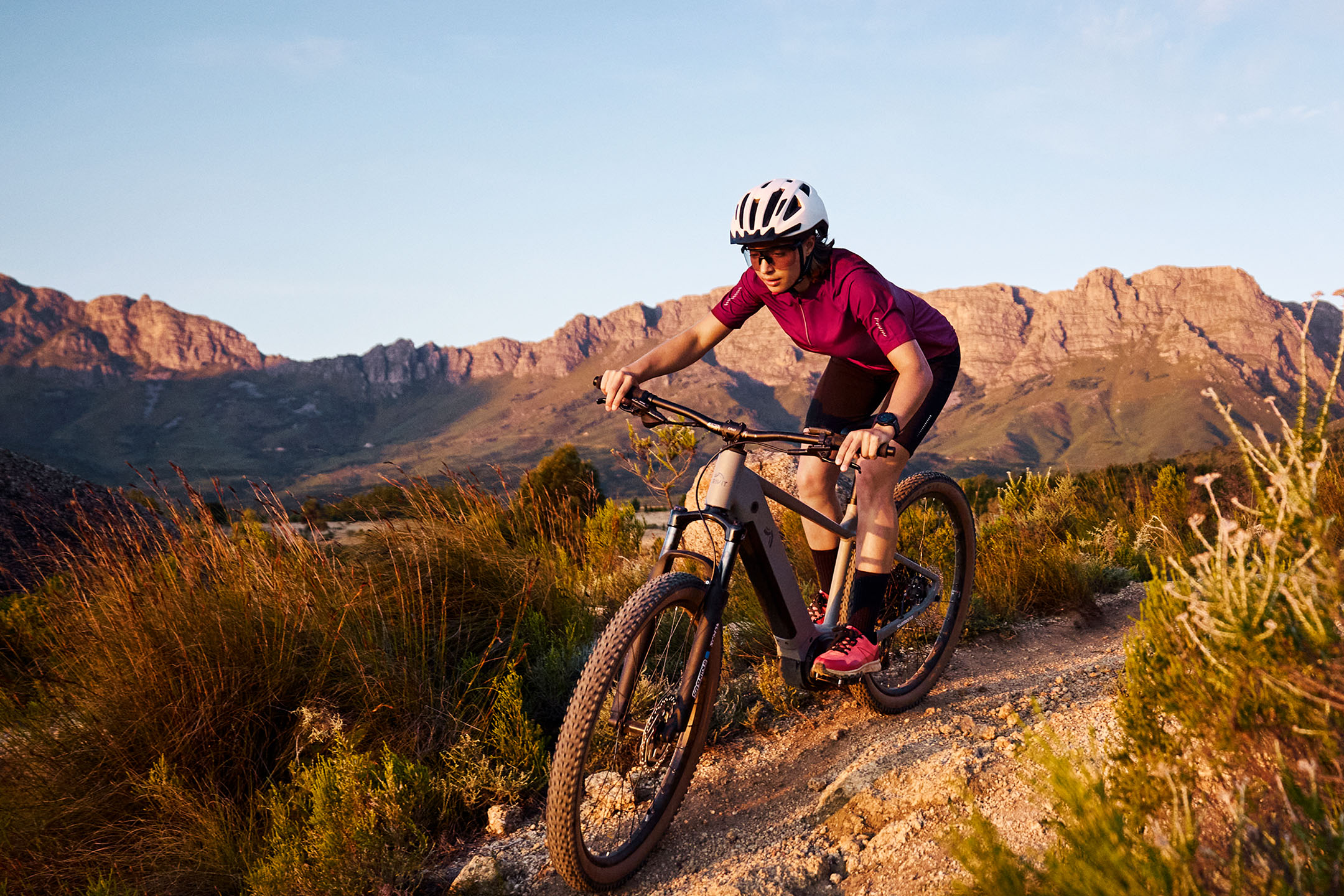 Frau fährt Mountainbike auf einem Feldweg mit Bergen im Hintergrund.
