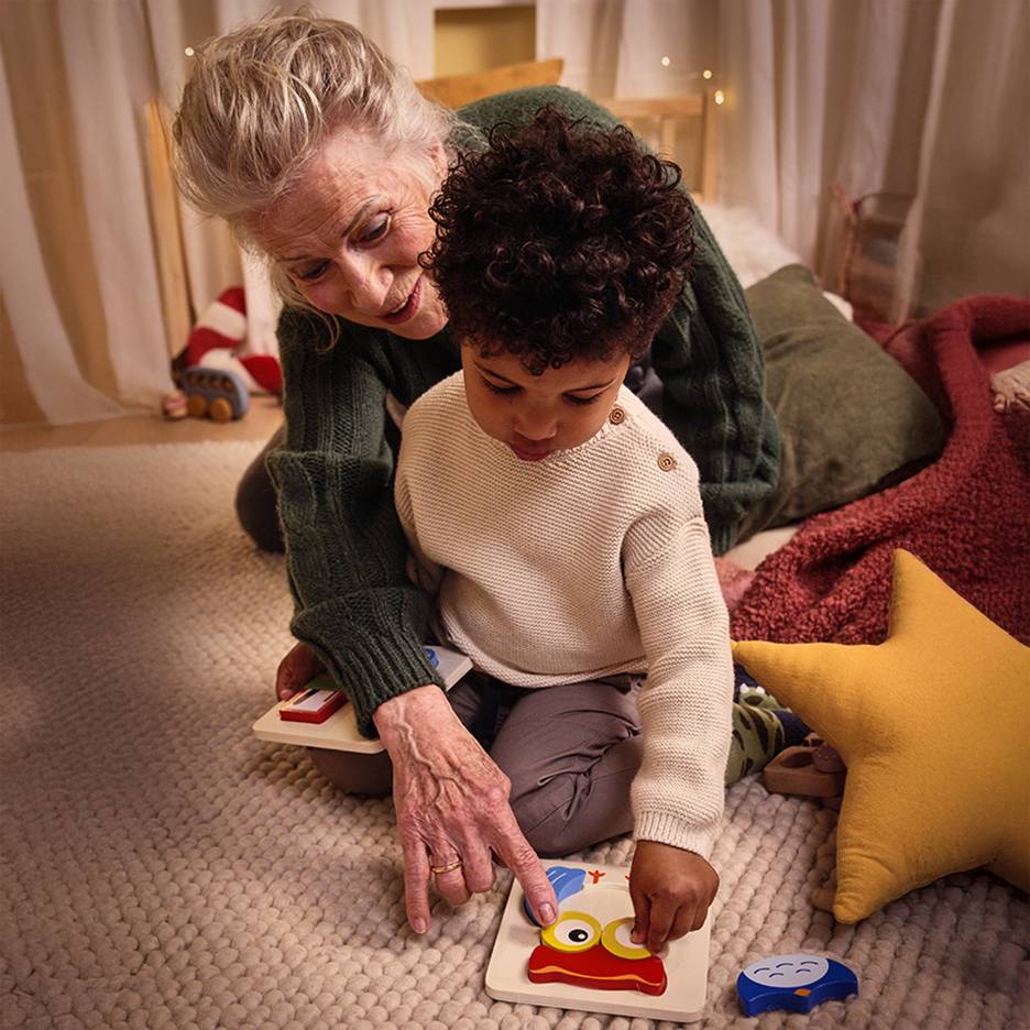 Großmutter und Kind spielen mit einem Holzpuzzle auf dem Teppich.