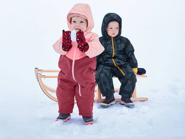 Zwei Kinder in Winteroveralls und Handschuhen auf einem Schlitten im Schnee.