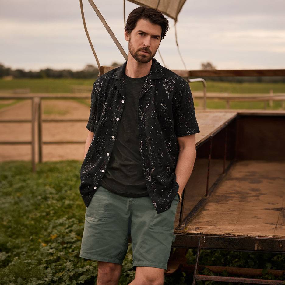 Mann in schwarzem gemustertem Hemd, T-Shirt und grünen Shorts, steht neben einem Anhänger.
