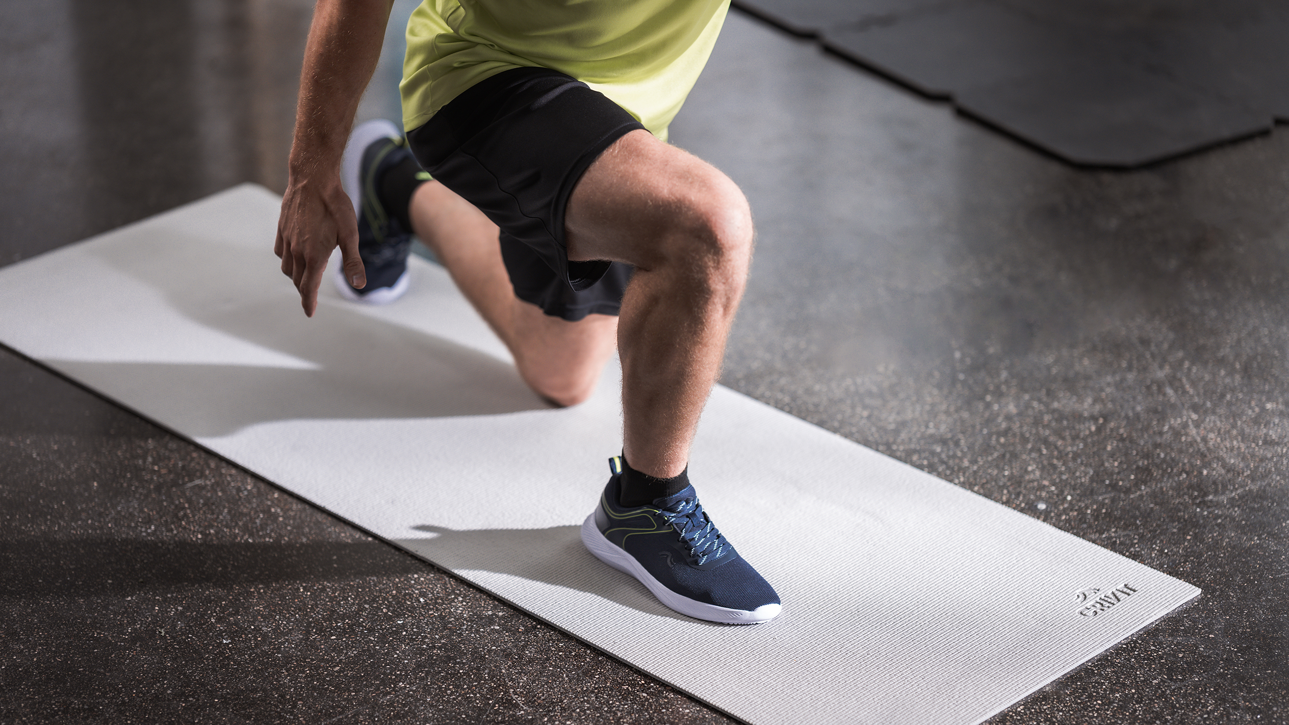 Mann in Sportkleidung macht Ausfallschritte auf einer hellgrauen Gymnastikmatte.