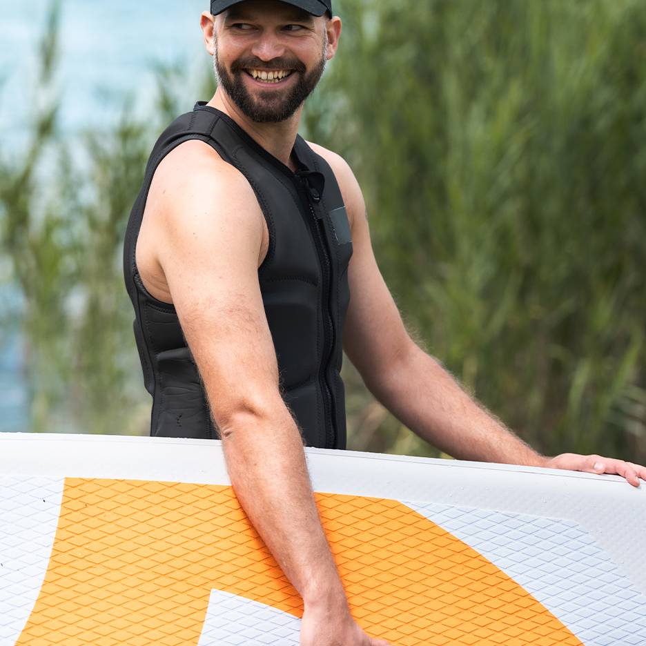 Lächelnder Mann in schwarzer Schwimmweste hält ein Stand-Up-Paddle-Board.