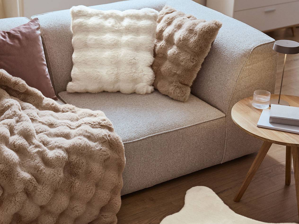 Sofa mit flauschigen Kissen und Decke, neben einem Holztisch mit Lampe.