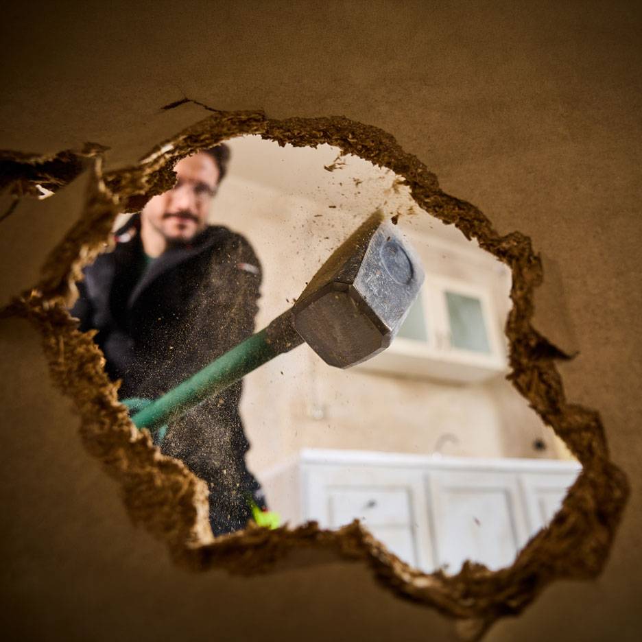 Mann mit Vorschlaghammer reißt Wand ein, durch ein Loch gesehen, mit Küchenschränken im Hintergrund.