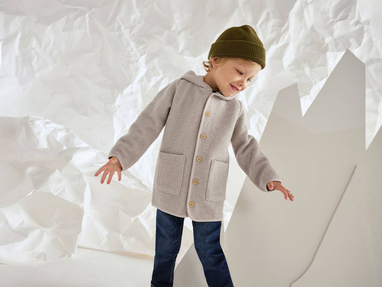 Junge in grauer Wolljacke und grüner Mütze, lächelnd vor zerknittertem Papierhintergrund.