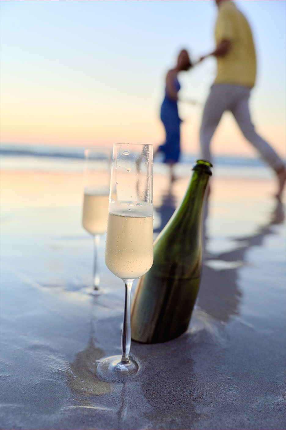 Sektgläser und Flasche am Strand bei Sonnenuntergang, Paar im Hintergrund.